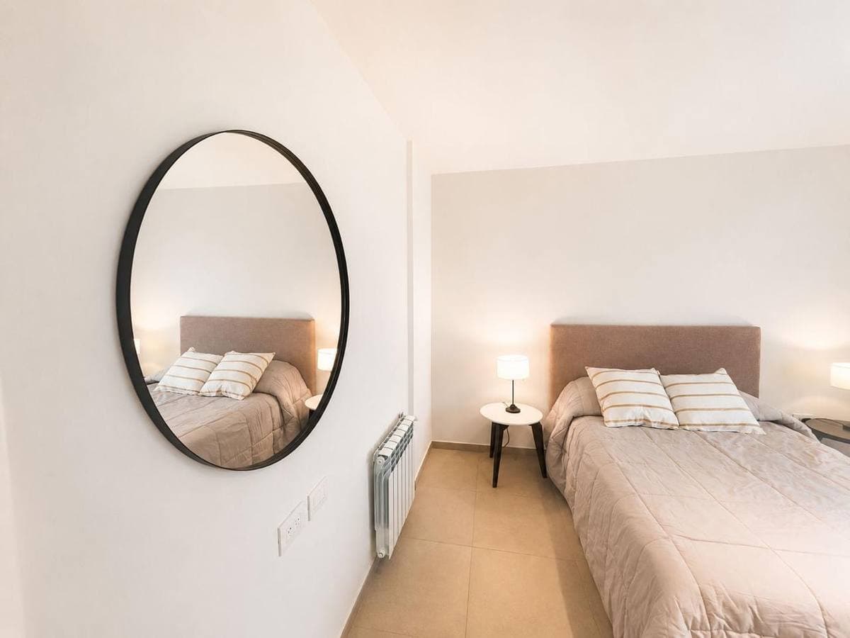 Dormitorio minimalista con un gran espejo ovalado negro que refleja una cama en tonos neutros.